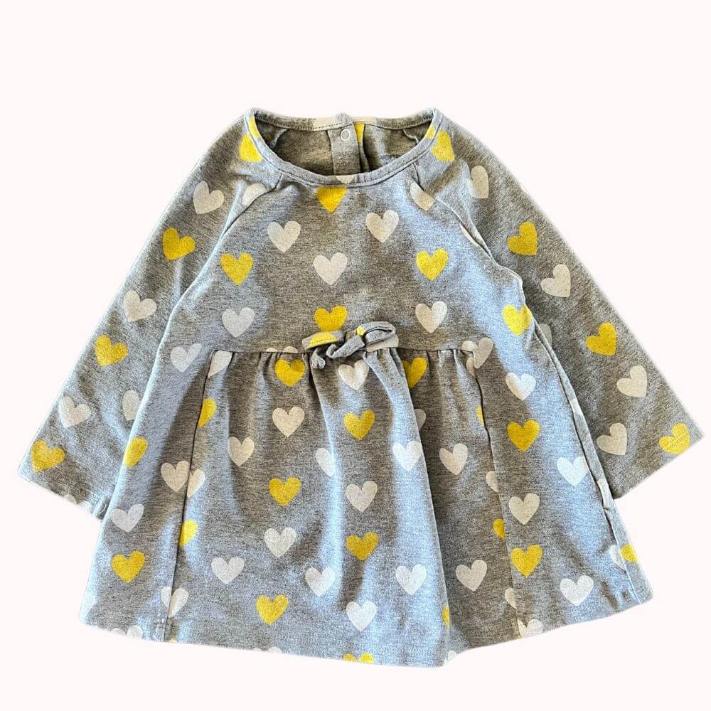 Robe bébé grise à manches longues, imprimé cœurs jaunes et blancs, coupe évasée avec taille froncée et petit nœud décoratif, fermeture boutonnée au dos.